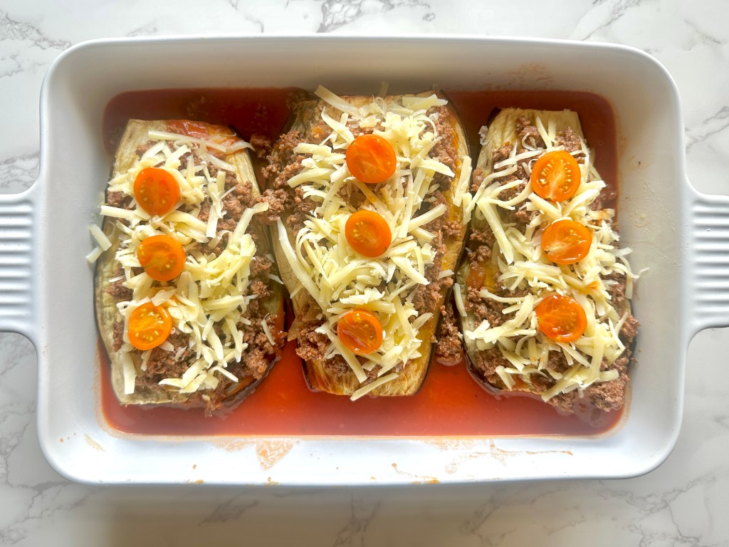 A baking dish with roasted eggplant, layered with a mixture of ground beef, onion, and spices, and topped with shredded cheddar cheese and grape tomatoes, sitting on a bed of marinara sauce