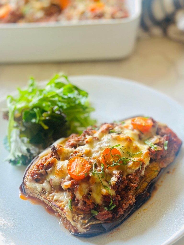 A serving plate of roasted eggplant casserole with ground beef, topped with melted cheddar and grape tomatoes, and served with a side salad