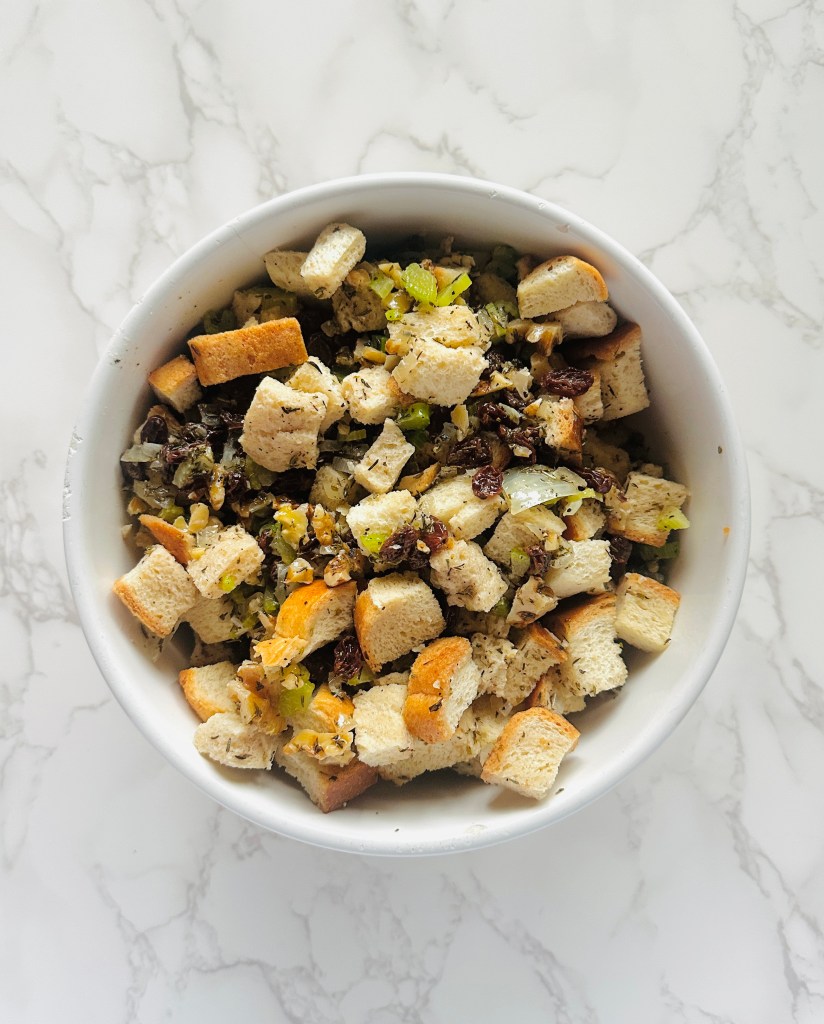 A large bowl of onion, celery, raisins, walnuts, and seasonings, topped with dried bread cubes