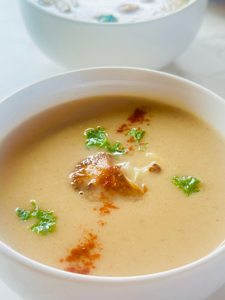 A serving bowl of roasted cauliflower soup, topped with cauliflower florets, chopped parsley, and ground allspice