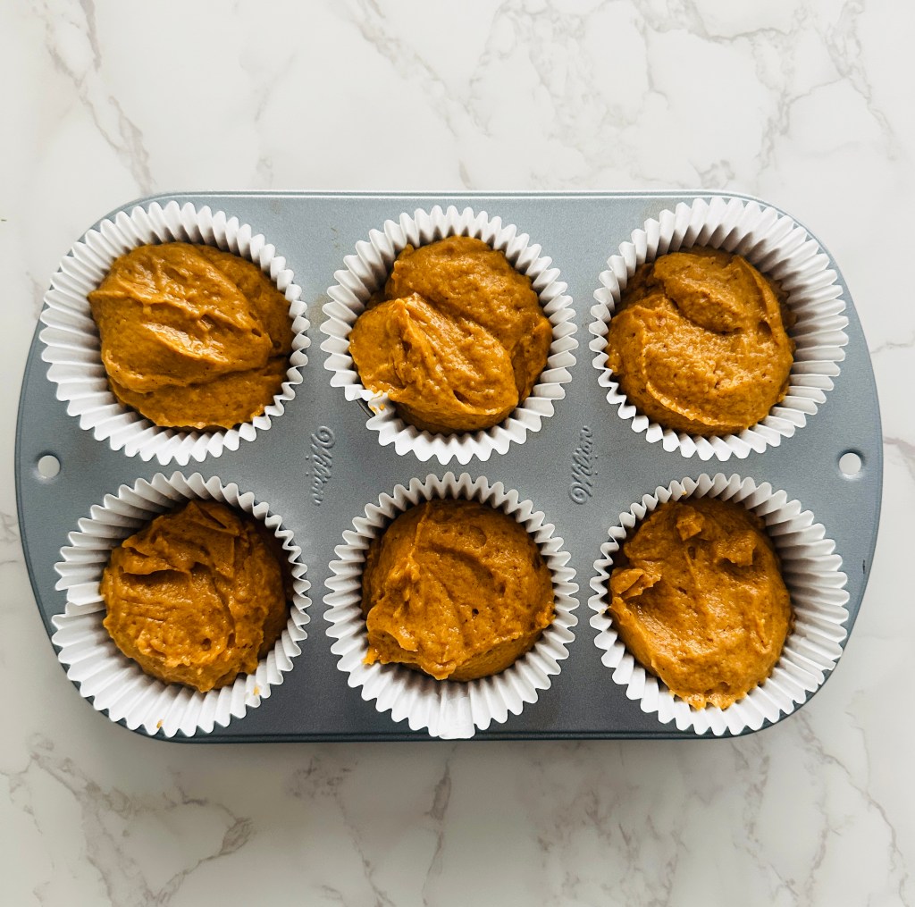 A 6-cup muffin tin with paper liners full of batter for pumpkin cupcakes