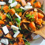 A serving plate of roasted sweet potato and arugula salad, combined with arugula, scallions, almonds, and feta, and topped with a cumin and lime dressing, being scooped up with a wooden spoon