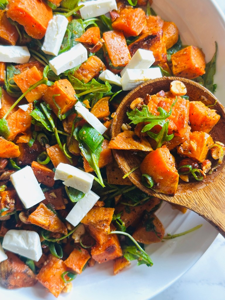 A serving plate of roasted sweet potato and arugula salad, combined with arugula, scallions, almonds, and feta, and topped with a cumin and lime dressing, being scooped up with a wooden spoon