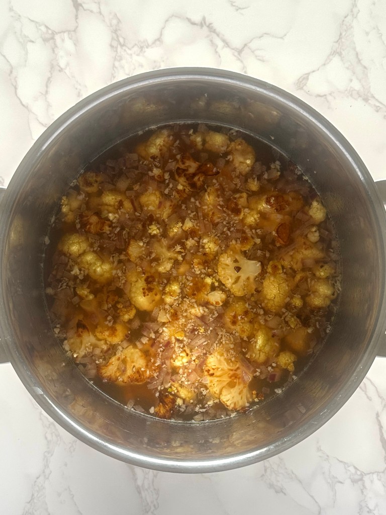 A stainless steel soup pot full of cooked onion, garlic, and vegetable broth mixed with roasted cauliflower