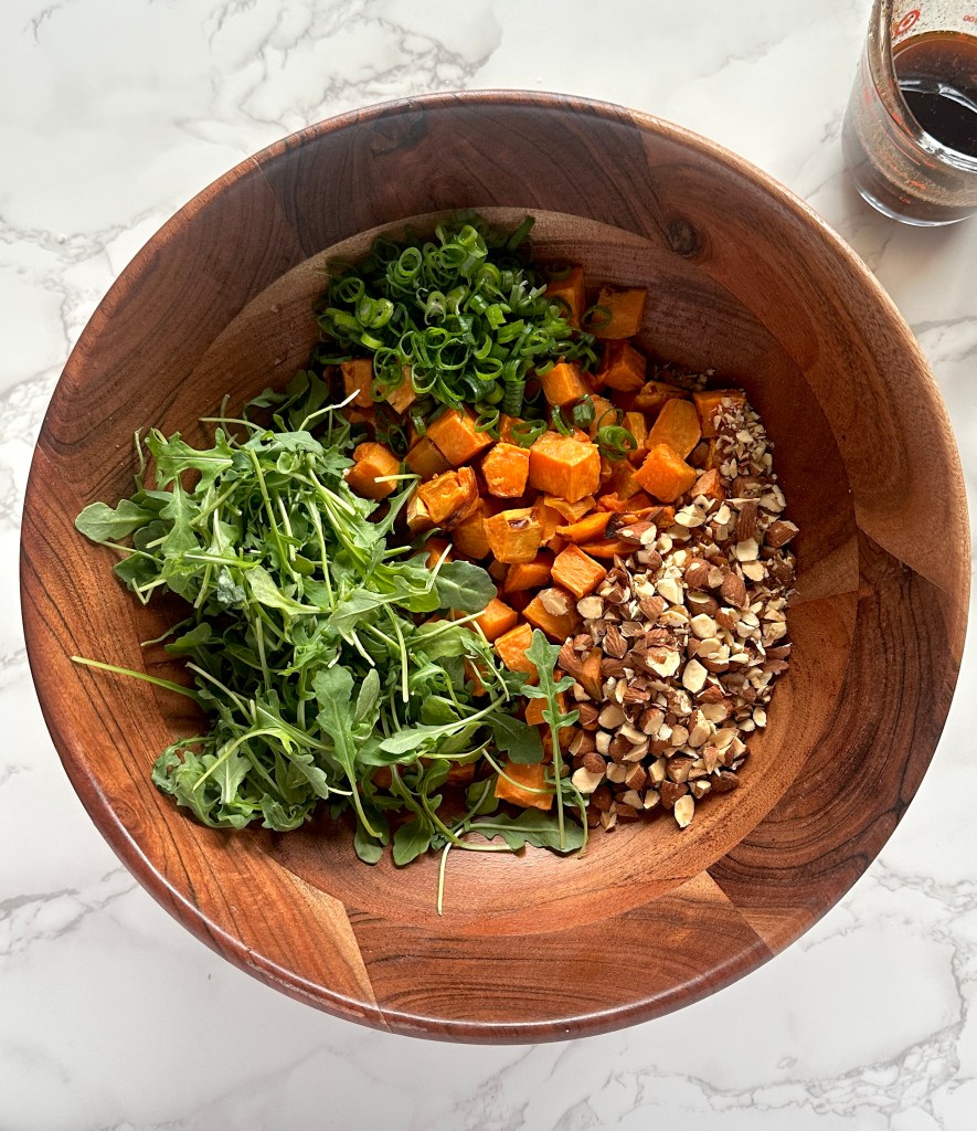 A large brown salad bowl full of arugula, sliced scallions, roasted sweet potatoes, and chopped almonds