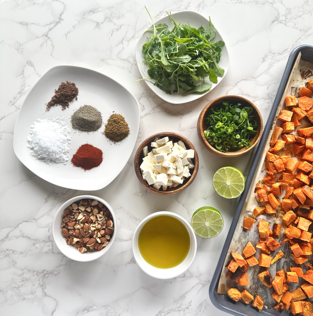 A photo of ingredients used to make sweet potato and arugula salad: roasted sweet potatoes, arugula, scallions, almonds, feta, and spices