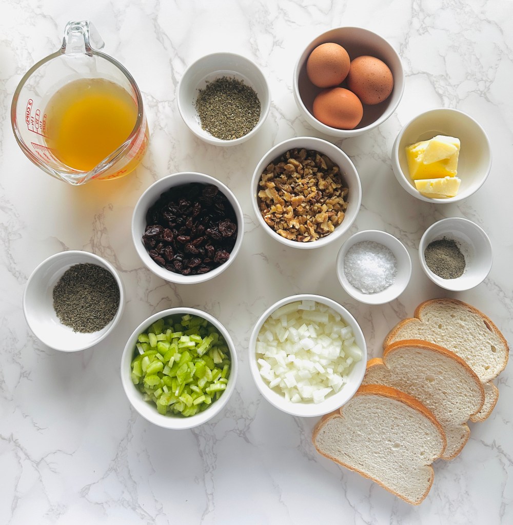 A photo of ingredients used to make Thanksgiving stuffing: white sandwich bread, onions, celery, raisins, walnuts, seasonings, butter, eggs, and chicken broth